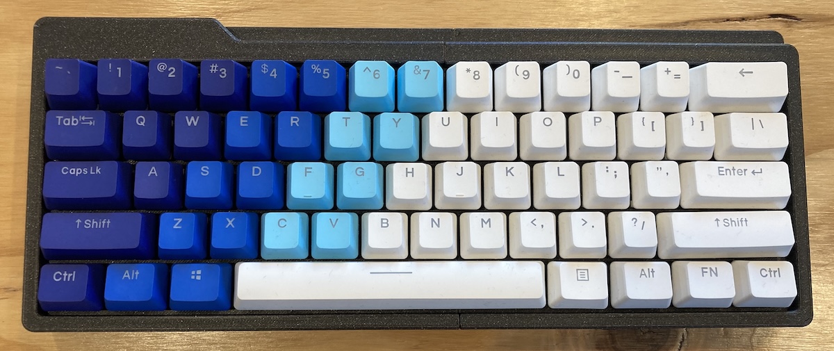 A small 60% keyboard. It has a black 3D printed case and the keycaps are dark blue, light blue and white in a gradientfrom left-to-right.